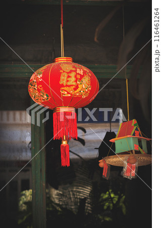 A traditional Chinese red lantern with gold designs and tassels hanging next to a hanging spirit house for the guardian spirit called neak ta in Khmer Buddhism belief 116461264