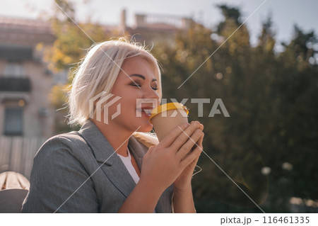 A blonde woman sits on a bench drinking coffee from a yellow cup. She is wearing a gray jacket and has her hair in a ponytail. The scene is peaceful and relaxing. A blonde woman sits on a bench drinking coffee from a yellow cup. She is wearing a gray jacket and has her hair in a ponytail. The scene is peaceful and relaxing. 116461335
