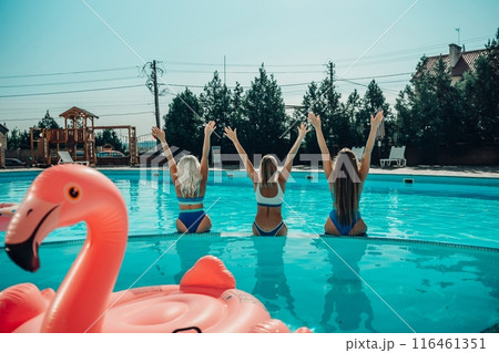 Three women are in a pool, one of them is sitting on a pink float. Scene is fun and lighthearted. 116461351