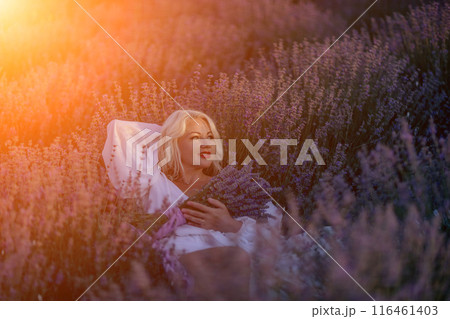 Blonde woman poses in lavender field at sunset. Happy woman in white dress holds lavender bouquet. Aromatherapy concept, lavender oil, photo session in lavender 116461403