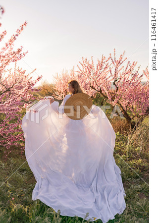 Woman blooming peach orchard. Against the backdrop of a picturesque peach orchard, a woman in a long white dress and hat enjoys a peaceful walk in the park, surrounded by the beauty of nature. Woman blooming peach orchard. Against the backdrop of a picturesque peach orchard, a woman in a long white dress and hat enjoys a peaceful walk in the park, surrounded by the beauty of nature. 116461417
