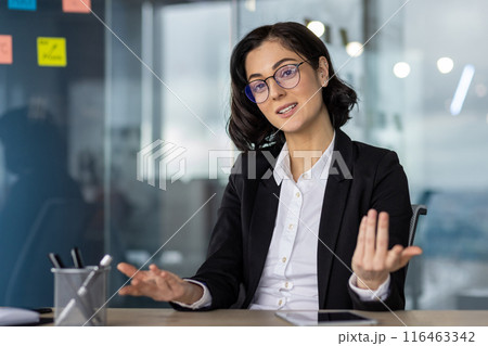 Businesswoman serious talking in video call meeting, explaining concepts or strategies. Professional setting with modern glass office background. Discussing issues or conducting interview. 116463342