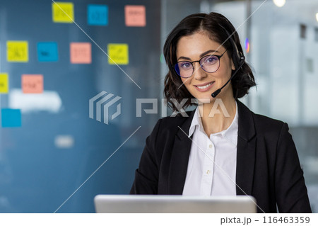 Smiling woman wearing headset working in modern office setting. Professional call center representative providing customer support, surrounded by colorful sticky notes. Smiling woman wearing headset working in modern office setting. Professional call center representative providing customer support, surrounded by colorful sticky notes. 116463359
