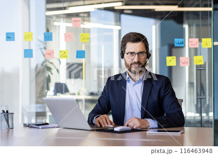 Portrait of successful businessman with headset phone, man in business suit smiling and looking at camera inside office, worker with laptop, helping buyers consumers online, support service. 116463948