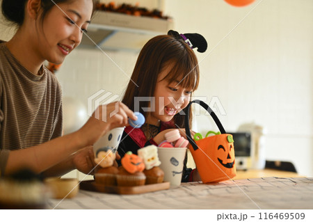 Cheerful little girl and mother decorating Halloween cupcakes with different monsters at kitchen table 116469509