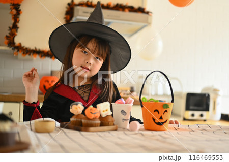 Portrait of cute little girl in witch costumes standing behind kitchen table with Halloween desserts 116469553