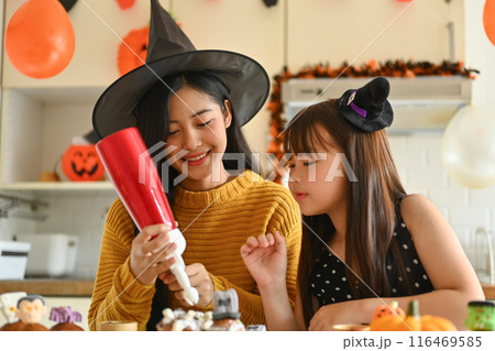 Happy smiling mother and daughter enjoying Halloween preparation at home, decorating cupcakes on table Happy smiling mother and daughter enjoying Halloween preparation at home, decorating cupcakes on table 116469585