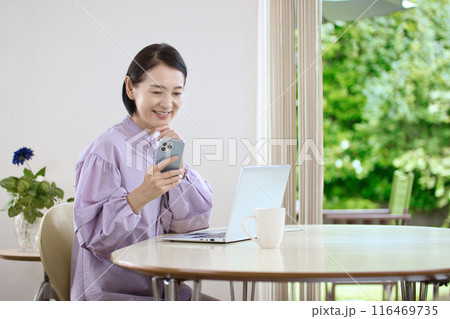 リビングでスマホを操作しながらパソコンを見るミドル女性 116469735