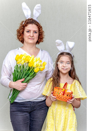 A woman with flowers and a girl with bunny ears and a basket of Easter eggs. The family celebrates Easter. 116471655