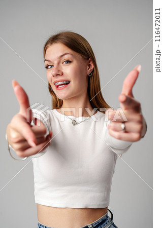 Smiling Young Woman With Braces Pointing Towards Camera in Studio Smiling Young Woman With Braces Pointing Towards Camera in Studio 116472011
