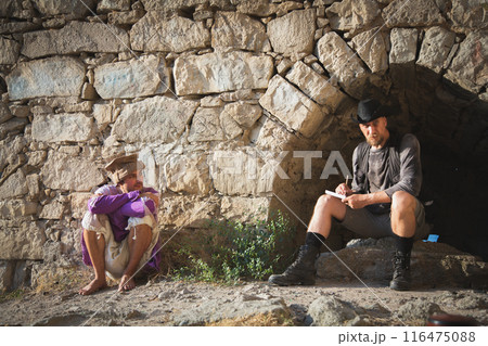 A man in traveler clothes and a hat writes against the background of an arched window of a fortress wall, near which a mercenary in a medieval Asian costume sits A man in traveler clothes and a hat writes against the background of an arched window of a fortress wall, near which a mercenary in a medieval Asian costume sits 116475088