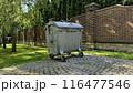 Metal trash can on wheels with lid against the backdrop of private household, trash container stands on the tiles near brick fence 116477546