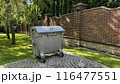 Trash on private territory against the backdrop of green lawn and brick fence, mobile trash container with lid near the house 116477551