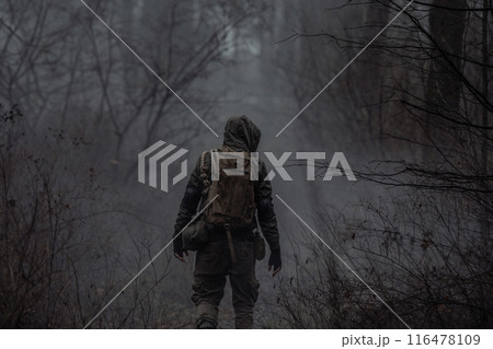 A man in camouflage in a post-apocalyptic, smoky, dark forest. Back view. 116478109