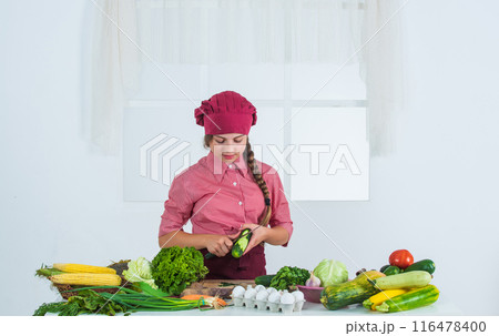 peel a cucumber. teen girl preparing meal. fresh raw vegetables. time to eat. happy child cooking in kitchen. healthy and organic food only. full of vitamins. kid wear chef uniform and hat peel a cucumber. teen girl preparing meal. fresh raw vegetables. time to eat. happy child cooking in kitchen. healthy and organic food only. full of vitamins. kid wear chef uniform and hat 116478400