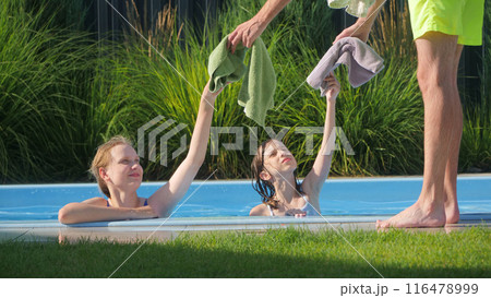 Two teenage girls in a pool reaching for towels handed by an adult, enjoying a sunny day. Two teenage girls in a pool reaching for towels handed by an adult, enjoying a sunny day. 116478999