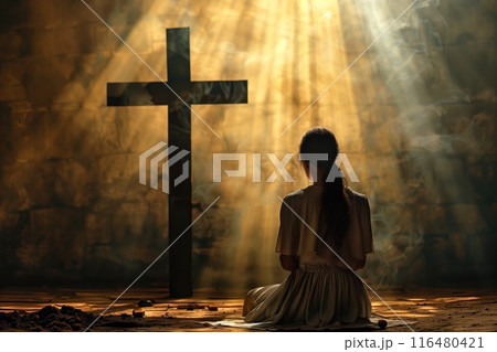 Woman kneels in front of large black cross, hands covering face. Stone wall and wooden floor provide rustic setting. Introspective moment captured in warm sunlight. 116480421