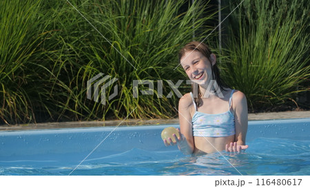 A teenage girl in a pool holding a ball and laughing, enjoying a sunny day. 116480617