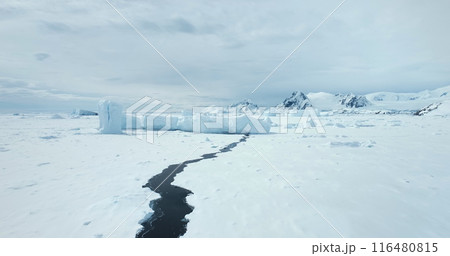 Fly over snow hill in Antarctica winter landscape. Frozen polar ocean with melted cracked river. Glacier and iceberg stuck in ice coast bay. Mountain range in background. Arctic travel and exploration 116480815