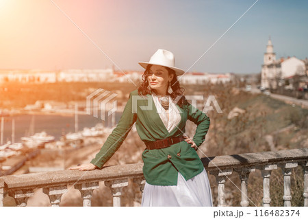 Woman walks around the city, lifestyle. Happy woman in a green jacket, white skirt and hat is sitting on a white fence with balusters overlooking the sea bay and the city. Woman walks around the city, lifestyle. Happy woman in a green jacket, white skirt and hat is sitting on a white fence with balusters overlooking the sea bay and the city. 116482374