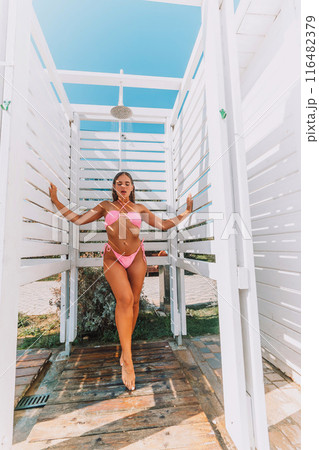 A woman in a bikini stands in front of a white shower stall. She is smiling and she is enjoying the moment. 116482379
