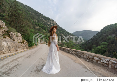 A woman in a white dress is standing on a road with a hat on 116482428