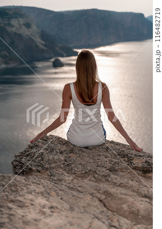 A woman sits on a rock overlooking the ocean. She is wearing a white tank top and white shorts. The scene is peaceful and serene, with the woman enjoying the view and the calming sound of the waves. A woman sits on a rock overlooking the ocean. She is wearing a white tank top and white shorts. The scene is peaceful and serene, with the woman enjoying the view and the calming sound of the waves. 116482719