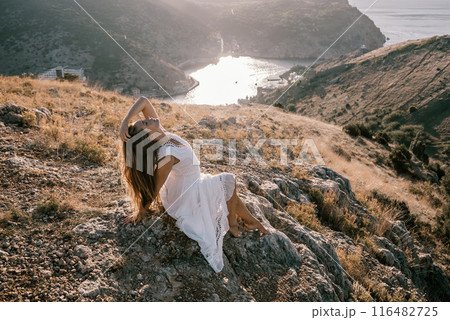 A woman in a white dress is sitting on a rock overlooking a body of water. She is enjoying the view and taking in the scenery. 116482725