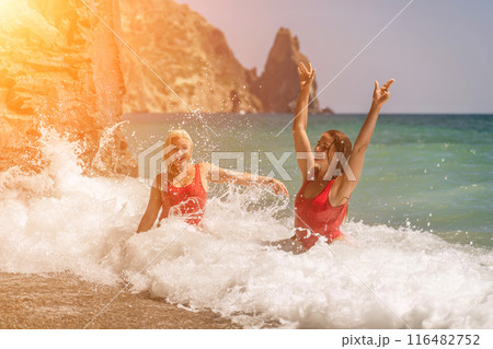 Women ocean play. Seaside, beach daytime, enjoying beach fun. Two women in red swimsuits enjoying themselves in the ocean waves and raising their hands up. Women ocean play. Seaside, beach daytime, enjoying beach fun. Two women in red swimsuits enjoying themselves in the ocean waves and raising their hands up. 116482752