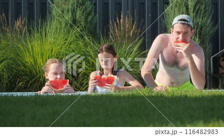 Two teenage girls and a man enjoying watermelon by the edge of a backyard pool. 116482983