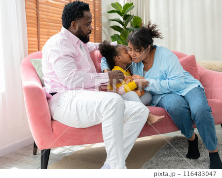 Happy family relaxing together on sofa at home. African little girl kissing her mom on cheek. . 116483047