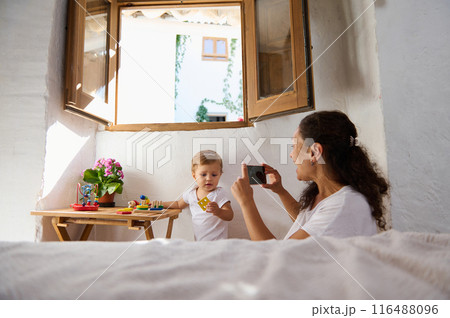 Mother taking photo of her toddler playing with toys near open window on a sunny day Mother taking photo of her toddler playing with toys near open window on a sunny day 116488096