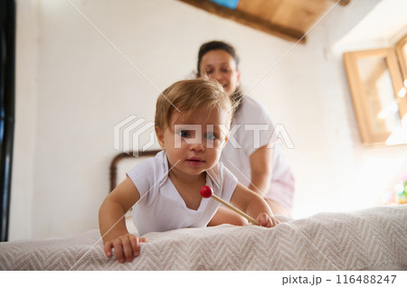Adorable baby crawling on bed with mother in background at home, cheerful and playful moment captured Adorable baby crawling on bed with mother in background at home, cheerful and playful moment captured 116488247