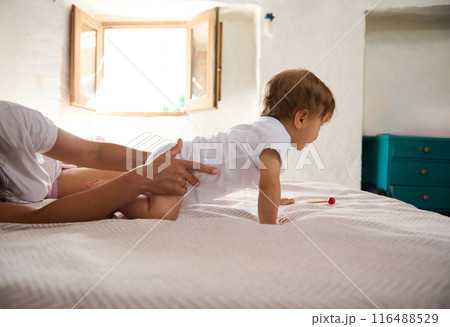 Parent supporting baby learning to crawl on bed in bright bedroom 116488529