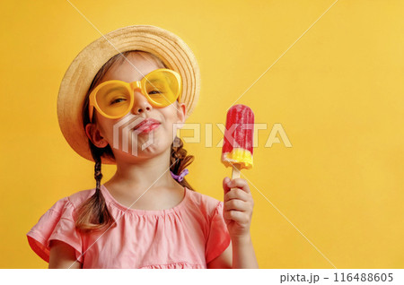 Cheerful little girl with fruit ice cream on yellow isolated background with copy space. Blonde female child with smirk on face in straw hat, sunglasses and pink top. She is holding a popsicle 116488605