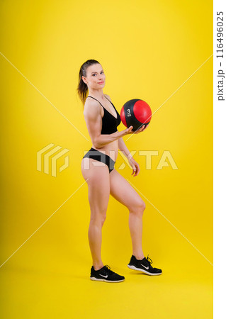 Sportive caucasian cross fit woman with fitness ball, look at camera. Studio dark yellow background 116496025