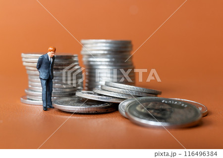 Miniature people A businessman in a suit stands with several stacks of silver coins Miniature people A businessman in a suit stands with several stacks of silver coins 116496384