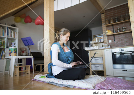 Newborn baby sleeping in baby sling, wrap, mother typing on laptop. Unconditional paternal love, mother's Day. 116499566