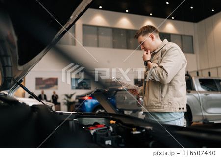 Low-angle view of pensive male customer holding papers, checking vehicle characteristics during choosing car in modern dealership center. Concept of choosing and buying new auto at showroom. Low-angle view of pensive male customer holding papers, checking vehicle characteristics during choosing car in modern dealership center. Concept of choosing and buying new auto at showroom. 116507430
