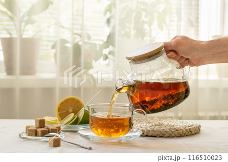 A hand pours brewed tea from glass teapot into a cup, with lemon slices and sugar cubes on the table near the window, cozy home environment 116510023
