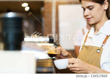 Professional Asian female barista pouring a shot of espresso into a coffee cup close up. Happy female barista making a cup of espresso coffee in a small coffee shop. Small business concept. Professional Asian female barista pouring a shot of espresso into a coffee cup close up. Happy female barista making a cup of espresso coffee in a small coffee shop. Small business concept. 116517669