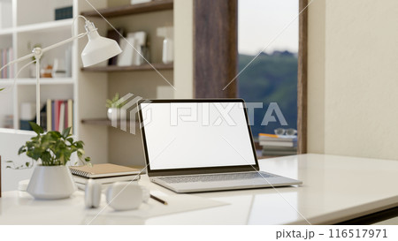 A close-up image of a laptop computer with a white-screen mockup on a white table in a modern room. 116517971