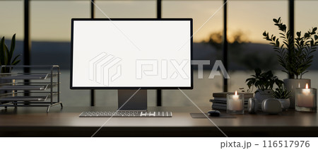 A computer with a white-screen mockup on a table against the window in a modern dark office at dusk. 116517976