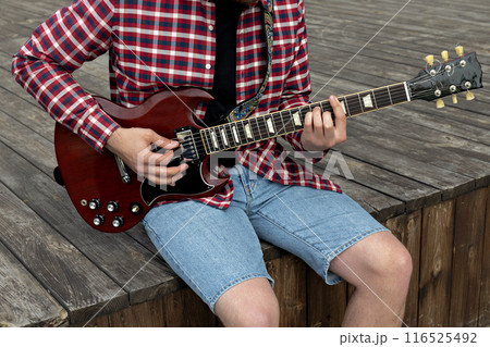 Stylish young guy playing guitar sitting on wooden stage Stylish young guy playing guitar sitting on wooden stage 116525492