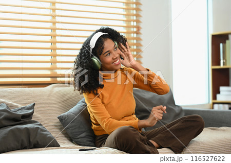 Cheerful young lady with curly hair sitting on couch, enjoying music through headphones 116527622