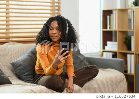 Relaxed young woman with curly hair wearing headphones listening to music, using mobile phone on sofa Relaxed young woman with curly hair wearing headphones listening to music, using mobile phone on sofa 116527634