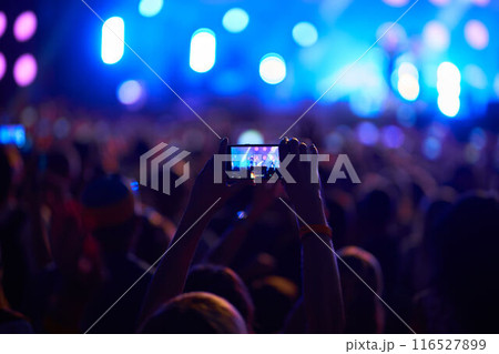 Crowd at music fest, backlit by stage lights, records live performance on smartphone. Excited fans in concert, capture artist in action, night event atmosphere. Diverse audience, outdoor venue. Crowd at music fest, backlit by stage lights, records live performance on smartphone. Excited fans in concert, capture artist in action, night event atmosphere. Diverse audience, outdoor venue. 116527899