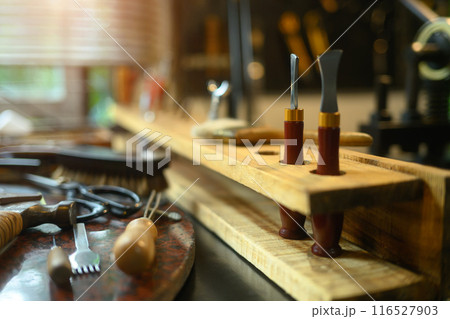 Various leather craft tools on wooden table in rustic workshop with natural light filtering through the blinds Various leather craft tools on wooden table in rustic workshop with natural light filtering through the blinds 116527903