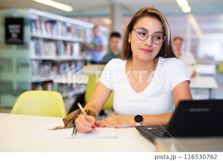 Focused Latin casual female in glasses writing in notepad and using laptop in the library 116530762