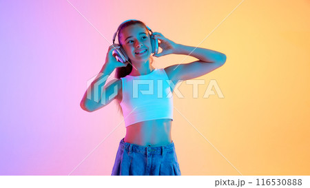 Portrait of beautiful teen girl in white top listening to music in headphones and smiling against gradient pink yellow background in neon light 116530888